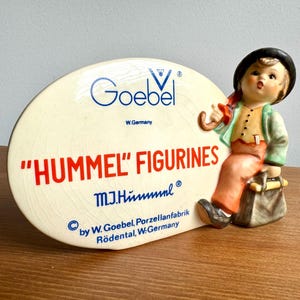 May include: A vintage Hummel figurine featuring a young boy with a black hat, green jacket, and orange pants, holding an umbrella. The figurine is next to a cream-colored sign with the Goebel logo and "HUMMEL" FIGURINES text.