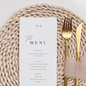 Peut inclure: Un menu blanc avec le texte "The Menu" et une liste de plats, placé sur un dessous de plat en osier. Une fourchette et un couteau dorés sont attachés avec un ruban rose clair. Le menu comprend les noms des plats et la date.