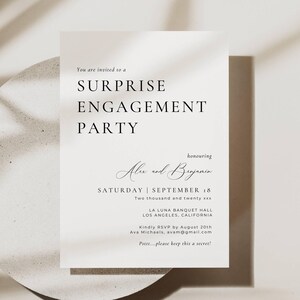 Könnte beinhalten: Eine weiße Einladungskarte für eine Überraschungs-Verlobungsfeier. Der Text lautet "SURPRISE ENGAGEMENT PARTY" mit den Namen "Alex und Benjamin" und dem Datum "SAMSTAG | 18. SEPTEMBER". Die Karte liegt auf einer runden, hellen Oberfläche.