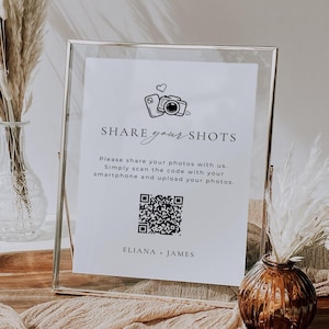 Puede incluir: Un marco de vidrio transparente contiene un letrero blanco con el texto "SHARE your SHOTS" y un código QR. El letrero también incluye los nombres "ELIANA + JAMES". El marco está sobre una mesa con jarrones decorativos y plantas secas.