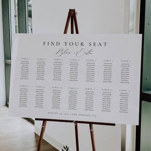 May include: A white seating plan displayed on a wooden easel. The text reads "FIND YOUR SEAT" above the names "Blair + Eastin". Guest names are listed by table number. The date "August 04, 2023" and location "Los Angeles, CA" are also included.
