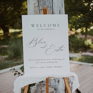 May include: A white welcome sign with black text announcing the wedding of Blair and Easton. The sign says "We're so glad you're here celebrating our love on our wedding day. July 10, 20XX | Paris, France."