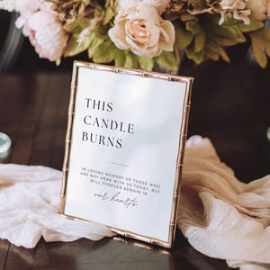 May include: A framed sign with the text "THIS CANDLE BURNS" in a gold bamboo-style frame. The sign includes additional text about remembering loved ones. The sign is placed on a dark wooden surface with a light pink fabric and floral arrangement in the background.
