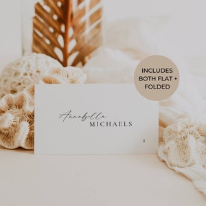 May include: White card with the name Annabella Michaels written in cursive script. The card is surrounded by white coral and a dried palm leaf. A brown circle with the text "Includes both flat + folded" is in the upper right corner.