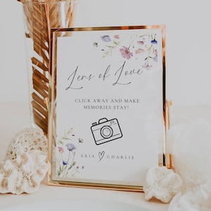 May include: A white sign with a gold frame and a floral design. The sign says "Lens of Love" and "Click away and make memories stay!" with a camera icon and the names "Aria & Charlie" at the bottom.