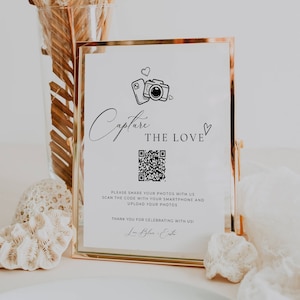 May include: A gold-framed sign with a white background and black text that reads "Capture the Love". The sign also features a QR code and a graphic of a camera and a smartphone. The text below the QR code reads "Please share your photos with us. Scan the code with your smartphone and upload your photos. Thank you for celebrating with us!"
