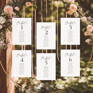 Può includere: Tabella dei posti a sedere per matrimoni con sei biglietti bianchi, ognuno con un numero di tavolo in elegante scrittura. I biglietti sono sospesi a un cerchio dorato adornato con fiori e verde. Nastri rosa pendono dal cerchio.