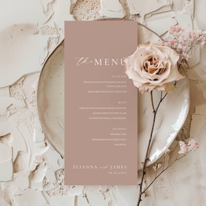 Può includere: Un biglietto del menu di nozze color rosa tenue con la scritta "The Menu" in corsivo. Il menu elenca antipasti, portate principali e dessert. Una grande rosa e piccoli rametti floreali adornano il biglietto, posto su un piatto bianco.