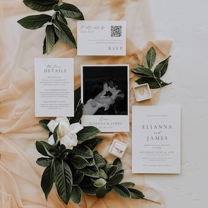 May include: Wedding invitation suite with a black and white photo, RSVP card, and details card. The suite includes a photo of a couple kissing, and the names ELIANNA and JAMES are printed on the cards. Green leaves and a white flower are arranged around the cards.