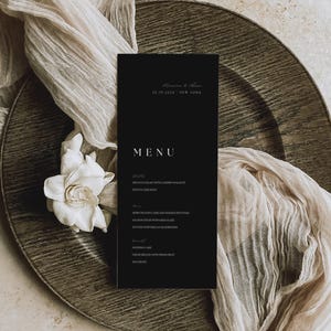 May include: A rectangular black menu card with white text featuring the heading MENU, followed by sections for starters, mains, and desserts, including the date 10.19.2024 and location New York. The card is accented by a white flower and fabric.