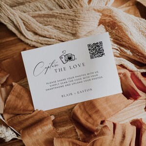 May include: A white rectangular card featuring black text and graphics. The card displays the words Capture THE LOVE, a camera icon, a QR code, and the text BLAIR + EASTON. Below the main title, it reads: PLEASE SHARE YOUR PHOTOS WITH US. SIMPLY SCAN THE CODE WITH YOUR SMARTPHONE AND UPLOAD YOUR PHOTOS.