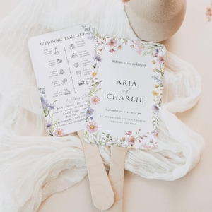 Könnte beinhalten: Zwei Hochzeitsfächer mit floralen Designs. Ein Fächer zeigt einen Hochzeitszeitplan mit Symbolen. Der andere Fächer sagt "Willkommen zur Hochzeit von ARIA und CHARLIE" mit Datum und Ort. Die Fächer haben Holzgriffe.