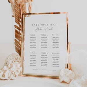 Puede incluir: Un letrero rectangular vertical con un marco de metal color cobre que muestra un plano de asientos de boda. El letrero blanco presenta el texto Take Your Seat, los nombres Blair + Easton, y enumera a los invitados para las mesas uno a seis con fecha y lugar.