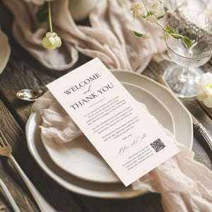 May include: A wedding reception place setting with a welcome card. The card reads "WELCOME and THANK YOU" in elegant script. The card is on a white plate with a soft pink fabric and a glass of water in the background.