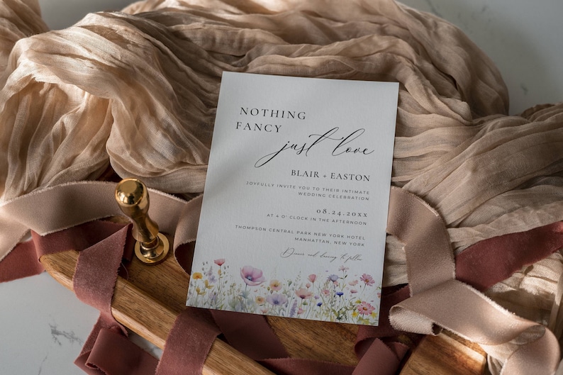 May include: A wedding invitation with a watercolor floral design. The invitation reads "Nothing Fancy Just Love" and includes the couple's names, Blair and Easton, the date of the wedding, 08.14.20XX, and the location, Thompson Central Park New York Hotel, Manhattan, New York.