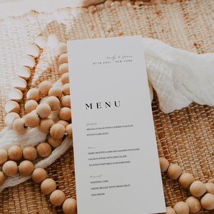 Op de afbeelding: Een witte menukaart voor een bruiloft met zwarte tekst. Het menu bevat voorgerechten, hoofdgerechten en desserts. De kaart ligt op een geweven mat met houten kralen.