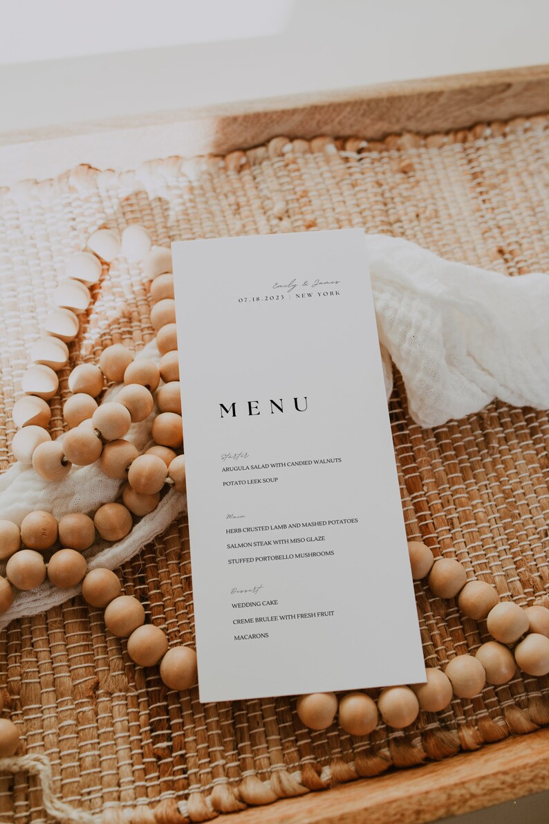 Op de afbeelding: Een witte menukaart voor een bruiloft met zwarte tekst. Het menu bevat voorgerechten, hoofdgerechten en desserts. De kaart ligt op een geweven mat met houten kralen.