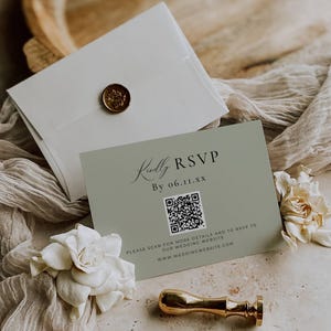 May include: A sage green RSVP card with the text "Kindly RSVP" and a QR code. The card is accompanied by a white envelope sealed with a gold wax stamp and a gold stamp handle. White flowers and fabric are in the background.