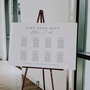 Puede incluir: Un plano de asientos de boda blanco sobre un caballete de madera. El texto "Find Your Seat" está sobre los nombres de los invitados, organizados por número de mesa. La fecha y el lugar están impresos en la parte inferior.