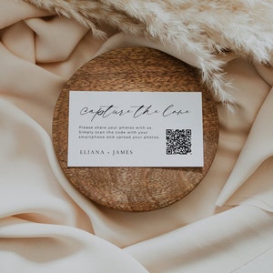 May include: White card with black text that reads "Capture the Cove" and a QR code. The text below the title reads "Please share your photos with us. Simply scan the code with your smartphone and upload your photos. ELIANA + JAMES". The card is on a wooden coaster.