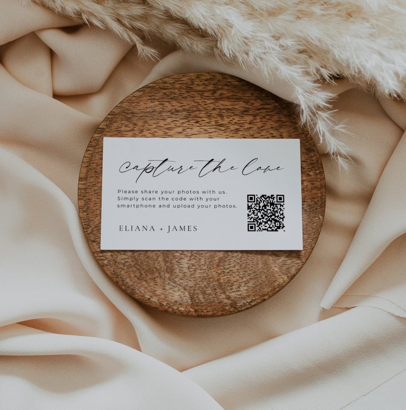May include: White card with black text that reads "Capture the Cove" and a QR code. The text below the title reads "Please share your photos with us. Simply scan the code with your smartphone and upload your photos. ELIANA + JAMES". The card is on a wooden coaster.