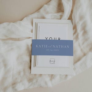 May include: A wedding invitation suite with a blue and white color scheme. The invitation features a white card with a blue band that says "Katie and Nathan" and the date "07.14.20XX".