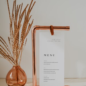 Op de afbeelding: Een koperen pijpstandaard met een witte kaart waarop een huwelijksmenu staat. Het menu bevat een salade, soep, lamsvlees, zalm, champignons, bruidstaart en cr&egrave;me br&ucirc;l&eacute;e. De standaard staat op een wit oppervlak met gedroogde bloemen in een roze vaas.