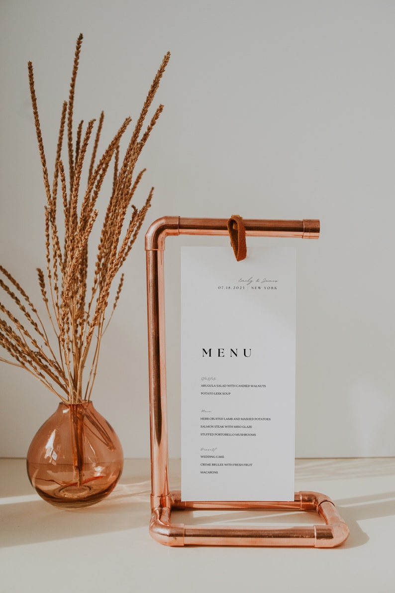 Op de afbeelding: Een koperen pijpstandaard met een witte kaart waarop een huwelijksmenu staat. Het menu bevat een salade, soep, lamsvlees, zalm, champignons, bruidstaart en cr&egrave;me br&ucirc;l&eacute;e. De standaard staat op een wit oppervlak met gedroogde bloemen in een roze vaas.