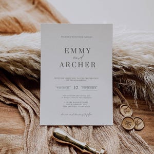 May include: A wedding invitation with the names "Emmy and Archer" printed in gray on a light-colored card. The invitation includes the date, time, and location of the ceremony. The card is on a textured surface with dried plants and wax seals.