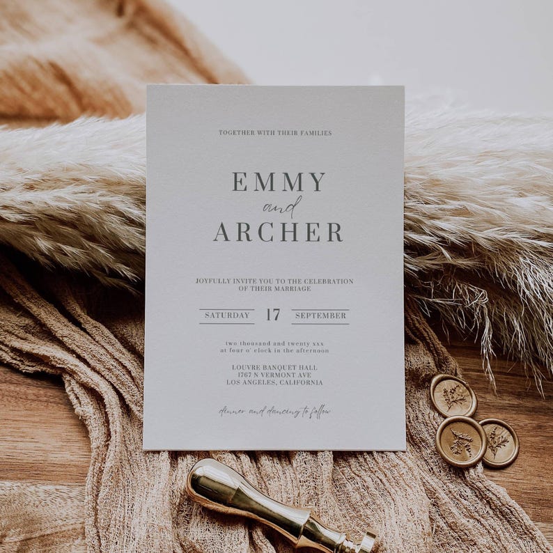 May include: A wedding invitation with the names "Emmy and Archer" printed in gray on a light-colored card. The invitation includes the date, time, and location of the ceremony. The card is on a textured surface with dried plants and wax seals.