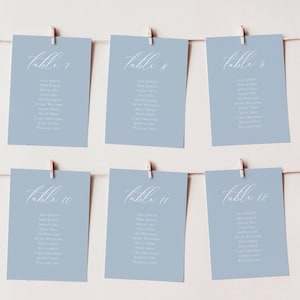 Puede incluir: Seis tarjetas de mesa azul claro con texto de caligrafía blanca, sujetas a una cuerda con pequeñas pinzas de madera. Cada tarjeta muestra un número de mesa y una lista de nombres, ideal para una boda o evento.