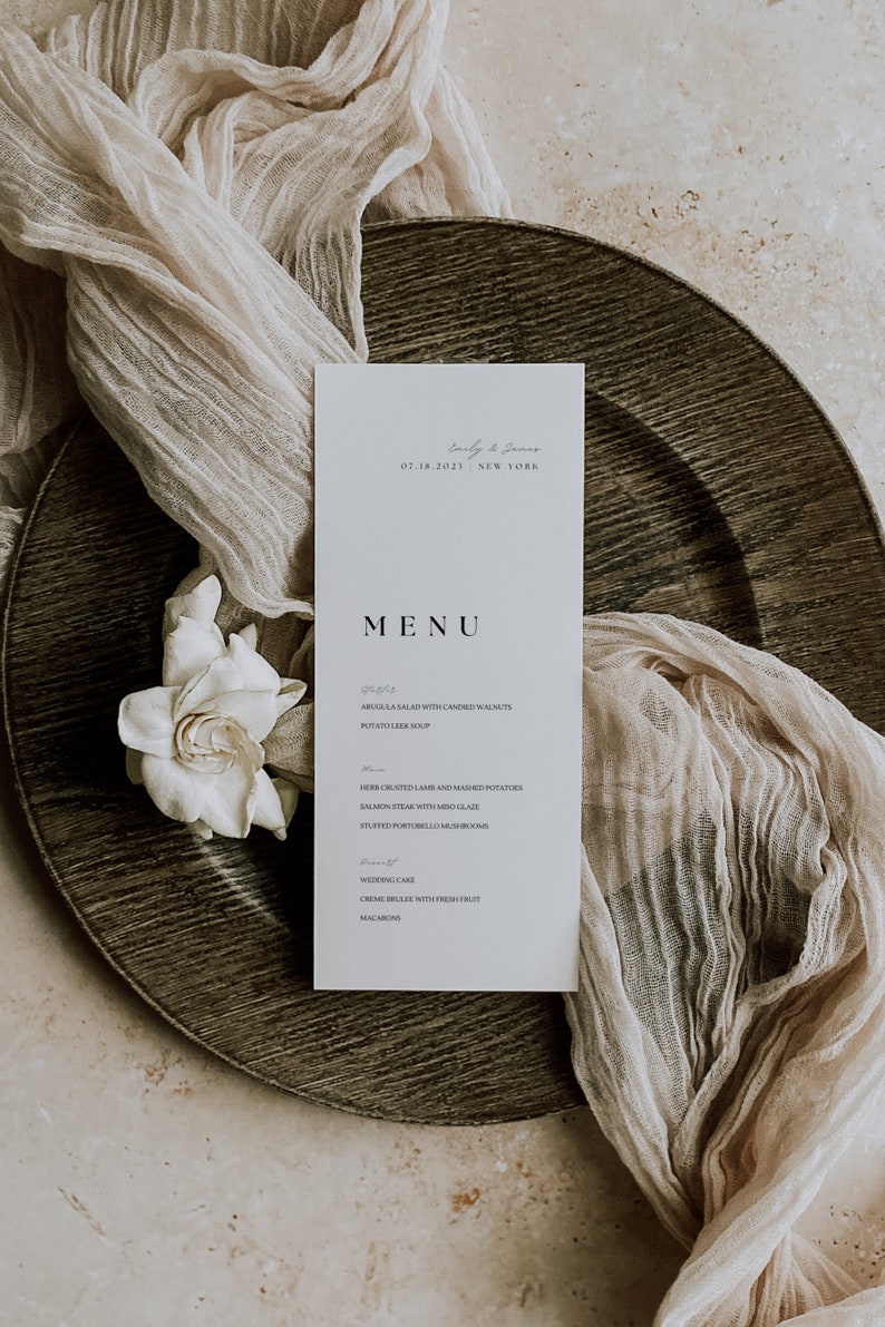 Op de afbeelding: Een wit menukaartje met zwarte tekst met de menukaart voor een huwelijksreceptie. Het menu bevat rucola salade met gekonfijte walnoten, aardappel-preisoep, kruidengekorst lamsvlees en gepureerde aardappelen, zalm met miso-glazuur, gevulde portobello's, bruidstaart, cr&egrave;me br&ucirc;l&eacute;e met vers fruit en macarons. Het kaartje ligt op een bruin houten bord met een witte doek.