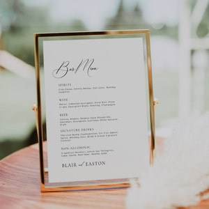 Könnte beinhalten: Ein goldfarbener Rahmen mit weißem Hintergrund und schwarzem Text listet die Bar-Speisekarte für einen Hochzeitsempfang auf. Die Speisekarte umfasst Spirituosen, Wein, Bier, Signature-Drinks und alkoholfreie Optionen. Das Schild enthält auch die Namen Blair und Easton.