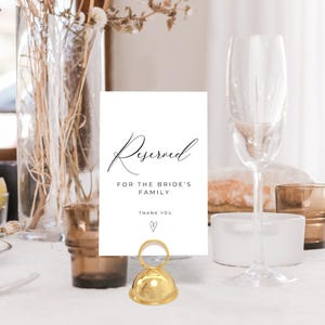 Könnte beinhalten: Eine weiße Karte mit dem Wort "Reserved" in eleganter Schrift, gehalten von einem goldfarbenen Metallhalter. Die Karte lautet auch "For the Bride's Family" und "Thank You" mit einem Herzsymbol. Ein Sektglas ist im Hintergrund.