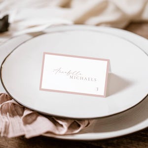 May include: A place card with a pale pink border and elegant script. The card reads "Annabella MICHAELS" with the number "3" in the bottom right corner. The card is set on a white plate with a folded napkin.