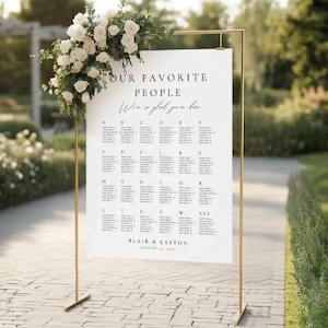 May include: A white wedding seating plan on a gold metal frame, decorated with white roses and foliage. The chart lists guest names alphabetically. Text reads "Our Favorite People, We're so glad you're here" and the names "Blair & Easton" with the date.