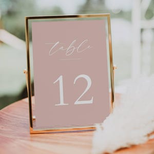 May include: A table number 12 sign in a gold frame. The sign has a blush pink background with the word "table" in cursive and the number 12 in a bold, clean font. The frame is resting on a wooden surface.