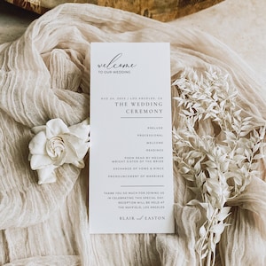 May include: A white wedding ceremony program with black text. The program details the wedding date, location, and order of service, including prelude, readings, and vows. It rests on cream fabric, with white floral decorations.