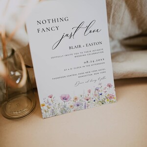 May include: A wedding invitation with a watercolor floral border. The text reads "Nothing Fancy, Just Love" with the names Blair and Easton, the date 08.24.20XX, and the location Thompson Central Park New York Hotel, Manhattan, New York.