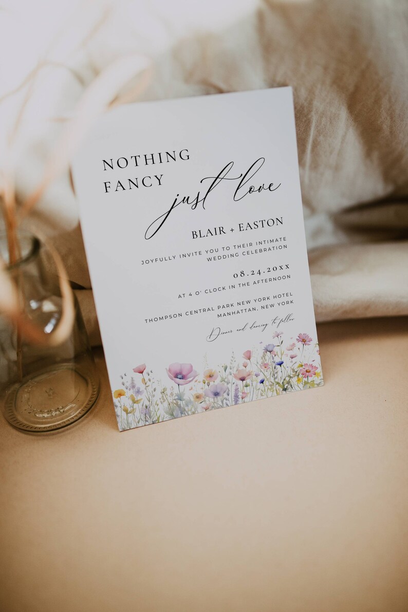 May include: A wedding invitation with a watercolor floral border. The text reads "Nothing Fancy, Just Love" with the names Blair and Easton, the date 08.24.20XX, and the location Thompson Central Park New York Hotel, Manhattan, New York.