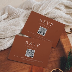 May include: Two terracotta RSVP cards with white text and QR codes. The cards say "RSVP" and "Please scan to RSVP to our wedding website." The date "08.12.23" is also displayed. The cards are on a wooden surface.