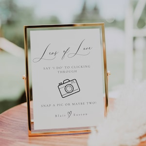 May include: A gold-framed sign with the text "Lens of Love" and a camera icon. The sign says "Say 'I do' to clicking through. Snap a pic or maybe two!" and is signed "Blair ♡ Easton".