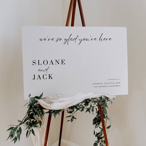 Könnte beinhalten: Ein weißes Schild auf einer Holzstaffelei mit dem Text "we're so glad you're here", "SLOANE and JACK" und dem Datum 24. August 2025 in Los Angeles, Kalifornien. Das Schild ist mit Grün und Stoff dekoriert.