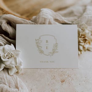 Puede incluir: Una tarjeta de agradecimiento blanca con un monograma floral dorado con las iniciales "B" y "E" y el texto "Gracias".