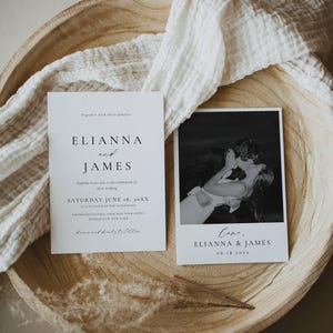 May include: Two white wedding invitation cards with black text. One card features the names "ELIANNA and JAMES" and wedding details, the other a black and white photo of a kissing couple. They rest on a wooden bowl with a white cloth.