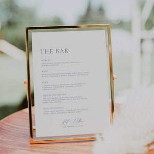 Könnte beinhalten: Ein goldfarbener Rahmen mit weißem Hintergrund listet die Bar-Speisekarte für einen Hochzeitsempfang auf. Die Speisekarte umfasst eine Auswahl an alkoholischen und alkoholfreien Getränken, darunter Spirituosen, Wein, Bier, Signature-Drinks und alkoholfreie Optionen. Das Schild steht auf einer Holzoberfläche mit einer weißen Federboa.