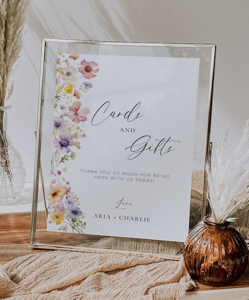 May include: A white sign with a floral border and the text "Cards and Gifts" in black script. The sign also includes the text "Thank you so much for being here with us today! Love, Aria + Charlie".