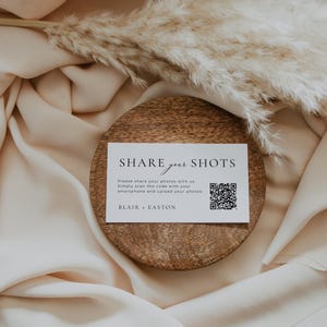 Pu&ograve; includere: Una carta rettangolare bianca con testo nero che recita SHARE your SHOTS, Please share your photos with us. Simply scan the code with your smartphone and upload your photos. BLAIR + EASTON, e un codice QR nero, appoggiata su un disco di legno.