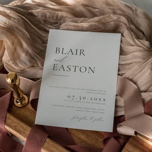 May include: A wedding invitation with the names "Blair and Easton" printed in black on a light gray card. The invitation includes the date, time, and location of the ceremony. A gold wax seal and ribbons are in the background.
