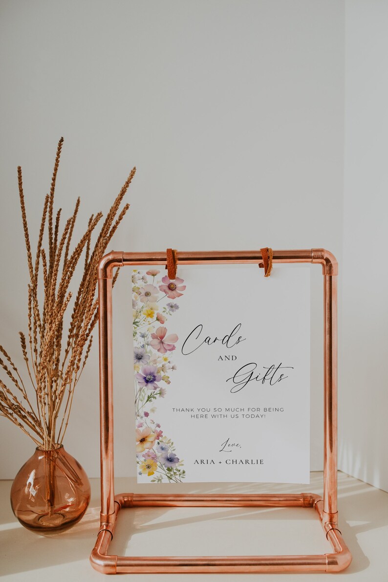 May include: A white sign with a floral border and the text "Cards and Gifts" with the text "Thank you so much for being here with us today! Love, Aria Charlie" below. The sign is displayed on a copper pipe stand.